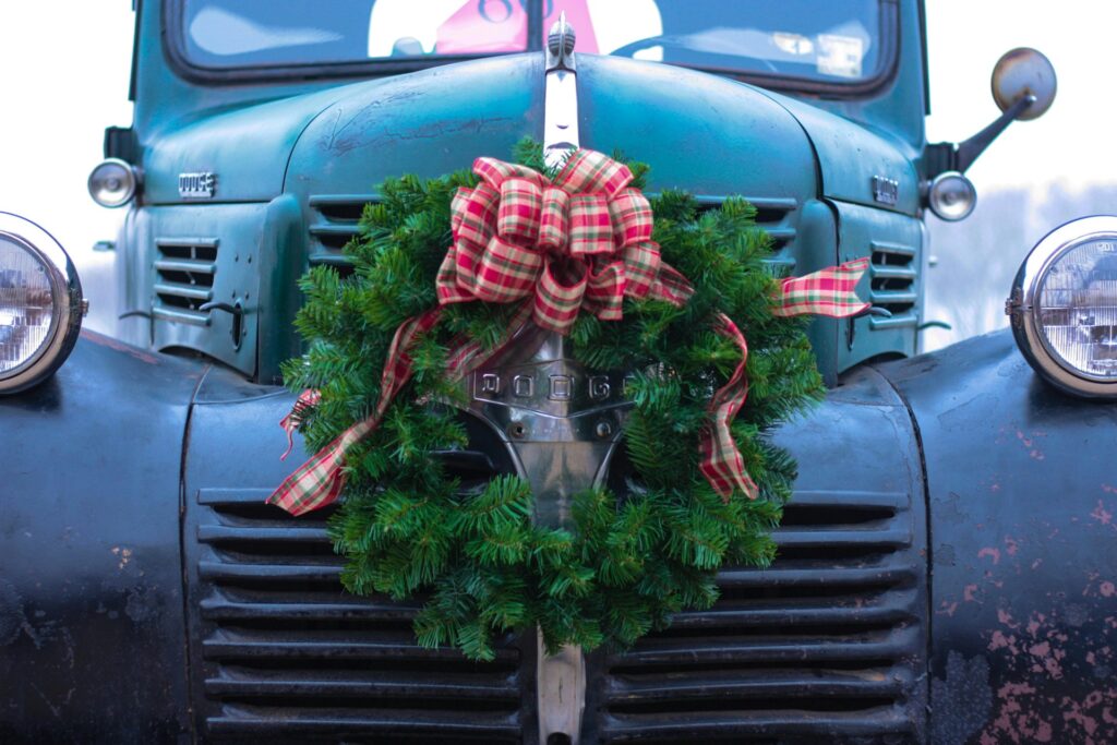 An early model of a Dodge car sits with a green wreath covered in a red plaid bow on the front of the hood. It evokes memories of the vintage Christmas aesthetic.