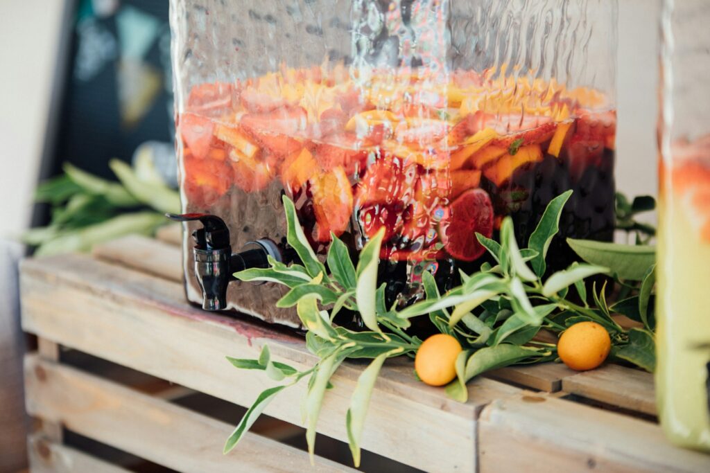 A large drink dispenser holding a pear sangria sits on a wooden crate surrounded by faux greenery and a few lemons