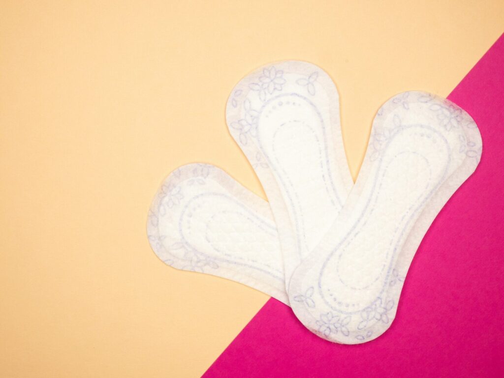 Three period panty liners sit unwrapped and flat against an orange and pink background. The pads are with with light purple flower designs.