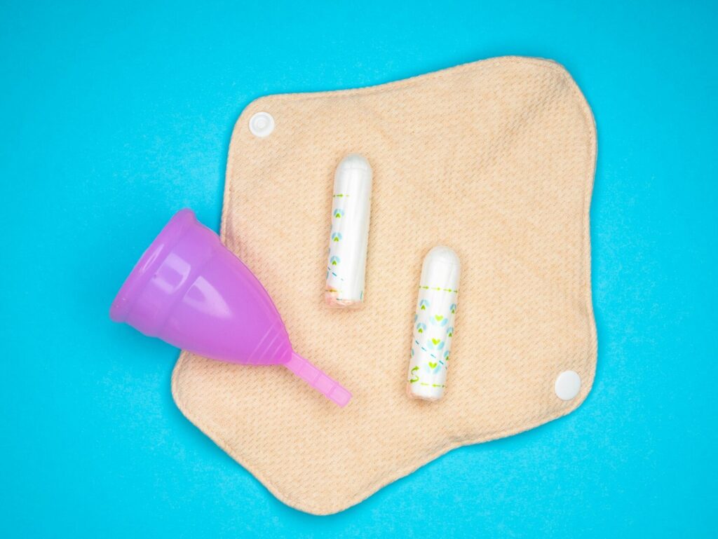 A homemade period pad with snap buttons sits open against a light blue background. A purple period cup and two stringless, applicator-less tampons sit on top of it.