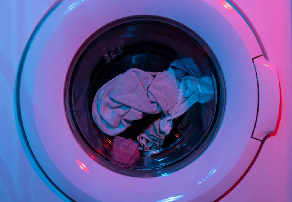 Clothes sit inside a washing machine that's white and has no brand on it. The exterior lighting around the machine is pink and blue.