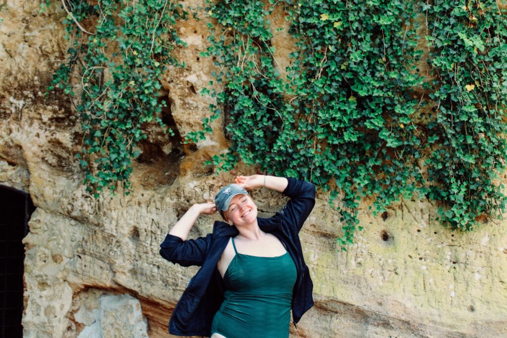 A woman in a green tank top and black jacket stands smiling with her hands on the baseball cap she wears. She stands in front of a rock structure covered in green vines.