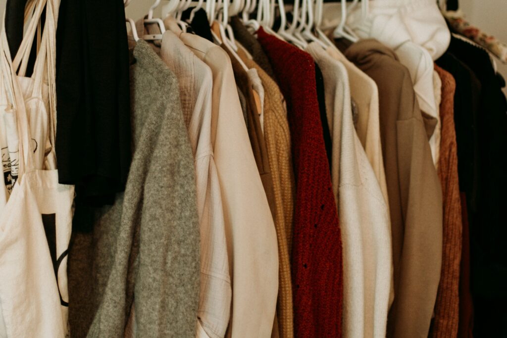 A row of sweaters on white hangers hangs from a clothing rack in a closet. The sweaters are all different colors, but most are in earth tones. 