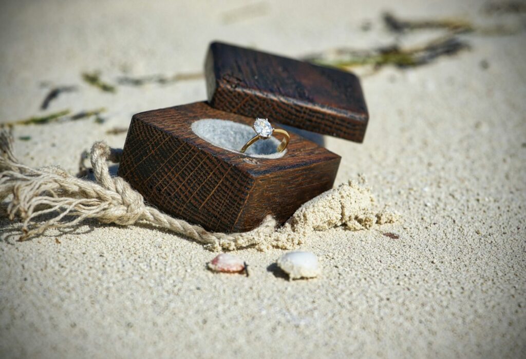 An oval diamond engagement ring sits in a velvet indent in a dark wooden engagement ring box. The lid is off and to the side. The box sits on a beach. There's a small rope next to it on the sand and two tiny shells the size of the ring.