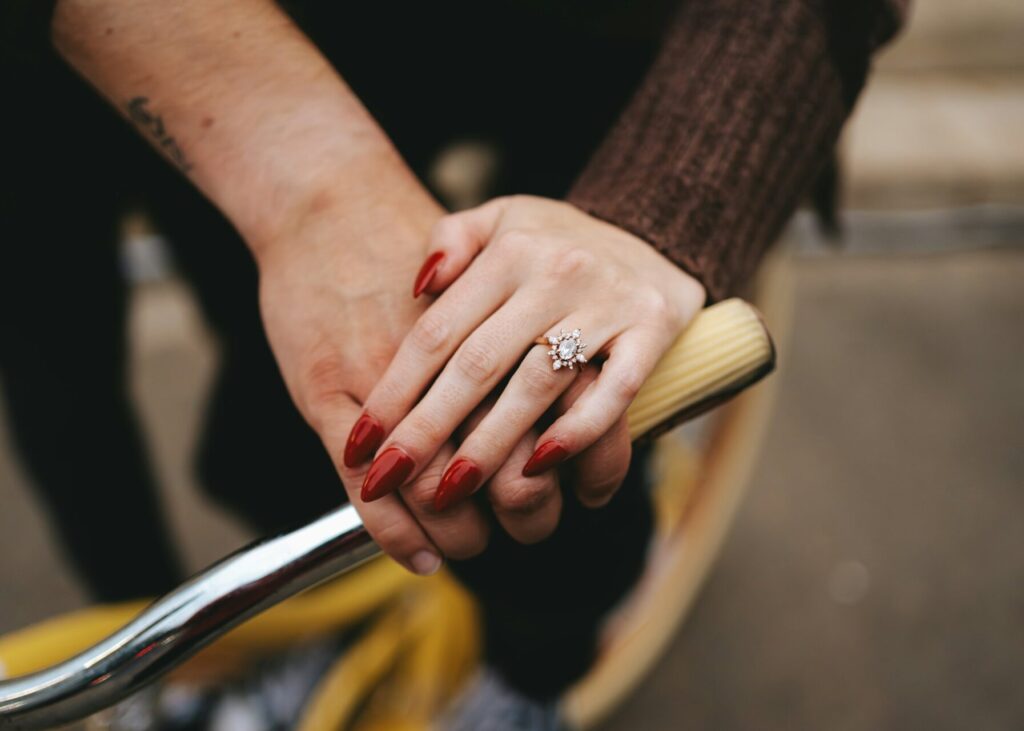 Two people have placed one hand each on the handle of an older metal bicycle. The woman's hand is on top of her partners. Her nails are long and red. She wears a sunburst engagement ring and a brown sweater. Her partner has a blurry tattoo on their arm.