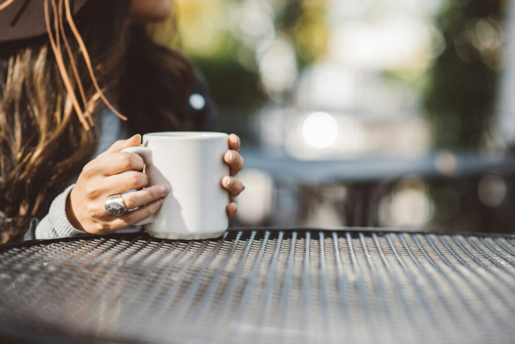 A woman sits at an outdoor black metal table holding a white mug filled with a drink.