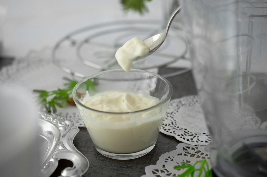 A small glass bowl filled with white yogurt sits on a gray table covered in other glass dishes and white doilies. A person spoons the yogurt out, potentially because it's one of the egg replacements for baking.