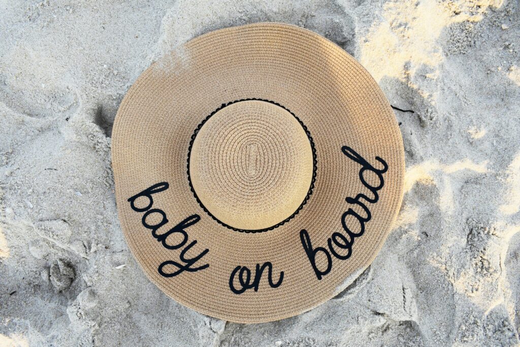 A woven sunhat sits on a sandy beach in the daylight. "Baby on board" is stitched around the rim.