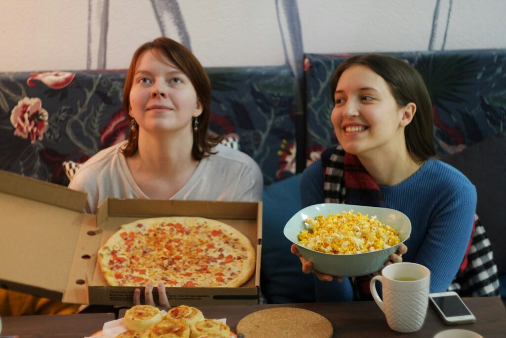 woman in white shirt sitting beside woman in red shirt eating pizza while watching shows like Tell Me Lies