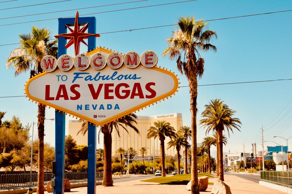 The Welcome to Fabulous Las Vegas Nevada sign stands under a blue sky. Palm trees surround it and the Mandalay Bay hotel is in the background.