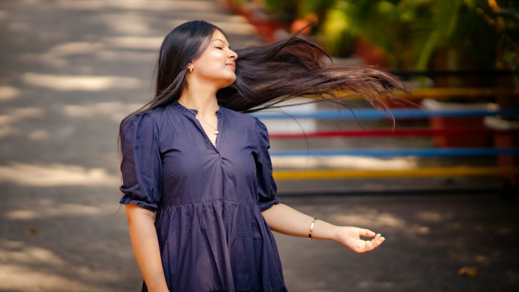 A woman in a navy dress tosses her long brown hair over one shoulder while walking along an outdoor path. She's likely participating in some of this year's hair trends.