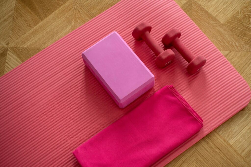 A pair of pink dumbbells sits on a pink yoga mat that's lying on a brown wooden floor. The mat also holds a pink exercise block and a dark pink towel that matches the pink Pilates princess aesthetic.